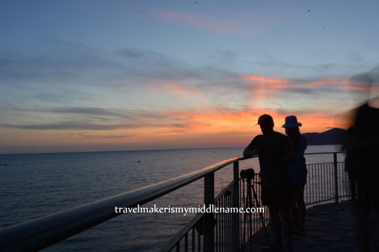 People looking at the sunset over the ocean on the lookout platform in Manarola. A few pink clouds are in the dark sky.