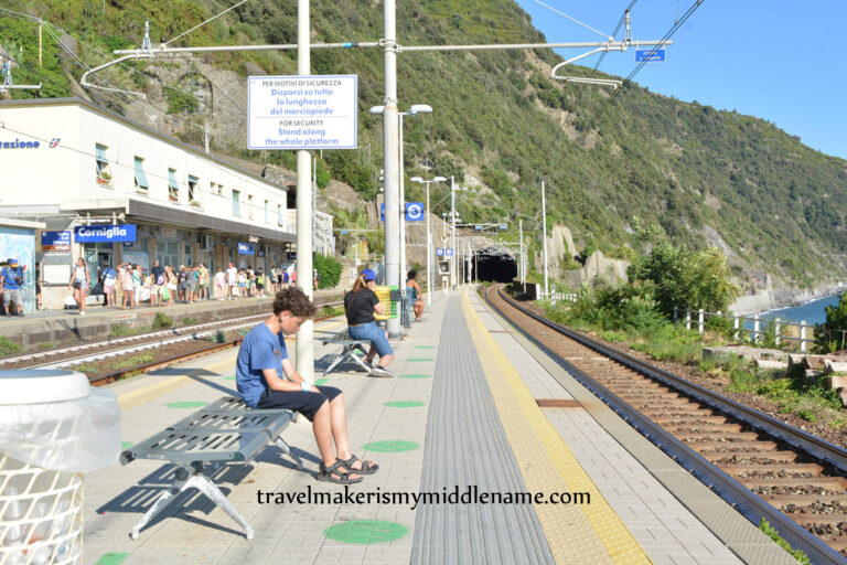 A person sits on a bench on the train platform at Corniglia train station, Italy. The train tunnel to the mountains is in the center of the image; railway tracks run along from the bottom right corner of the image to the tunnel, the green mountains is seen in the background, as well as a small portion of a cloudless blue summer sky in the top right corner.