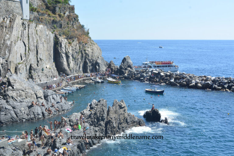 Ferry carrying passengers in the distance docks at Manarola, Italy. The left portion of the image are small cliffs, the right are the dark blue ocean waters with a ferry docked at a rocky bar. The light blue cloudless summer sky is in the distance.