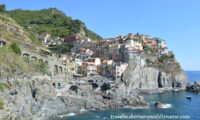 A wide shot of Riomaggiore in Cinque Terre in Italy showing the buildings on the cliffs. In the background are the green terraces for planting and a cloudless blue summer sky. Part of the dark blue ocean is seen in the foreground.