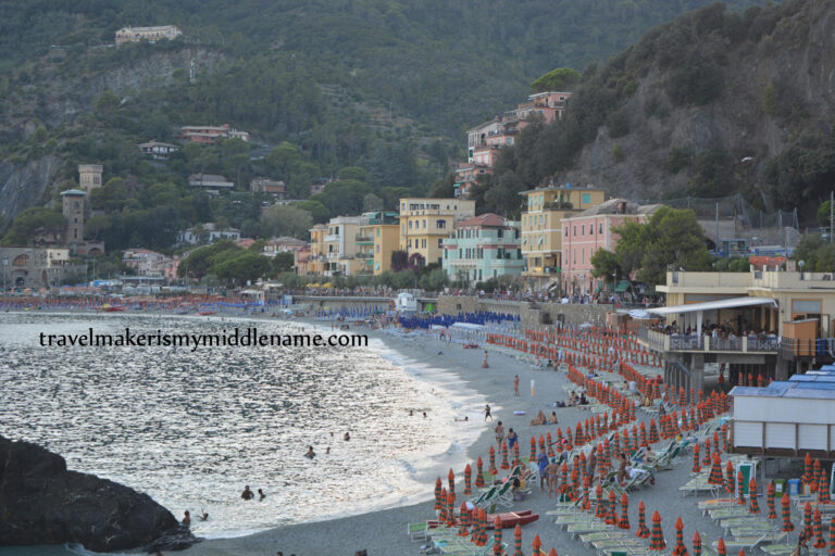 A greyish picture of Monterosso Beach, Italy which lies in the shadow of the cliffs at sunset. Many closed beach umbrellas line the entire length of the beach.