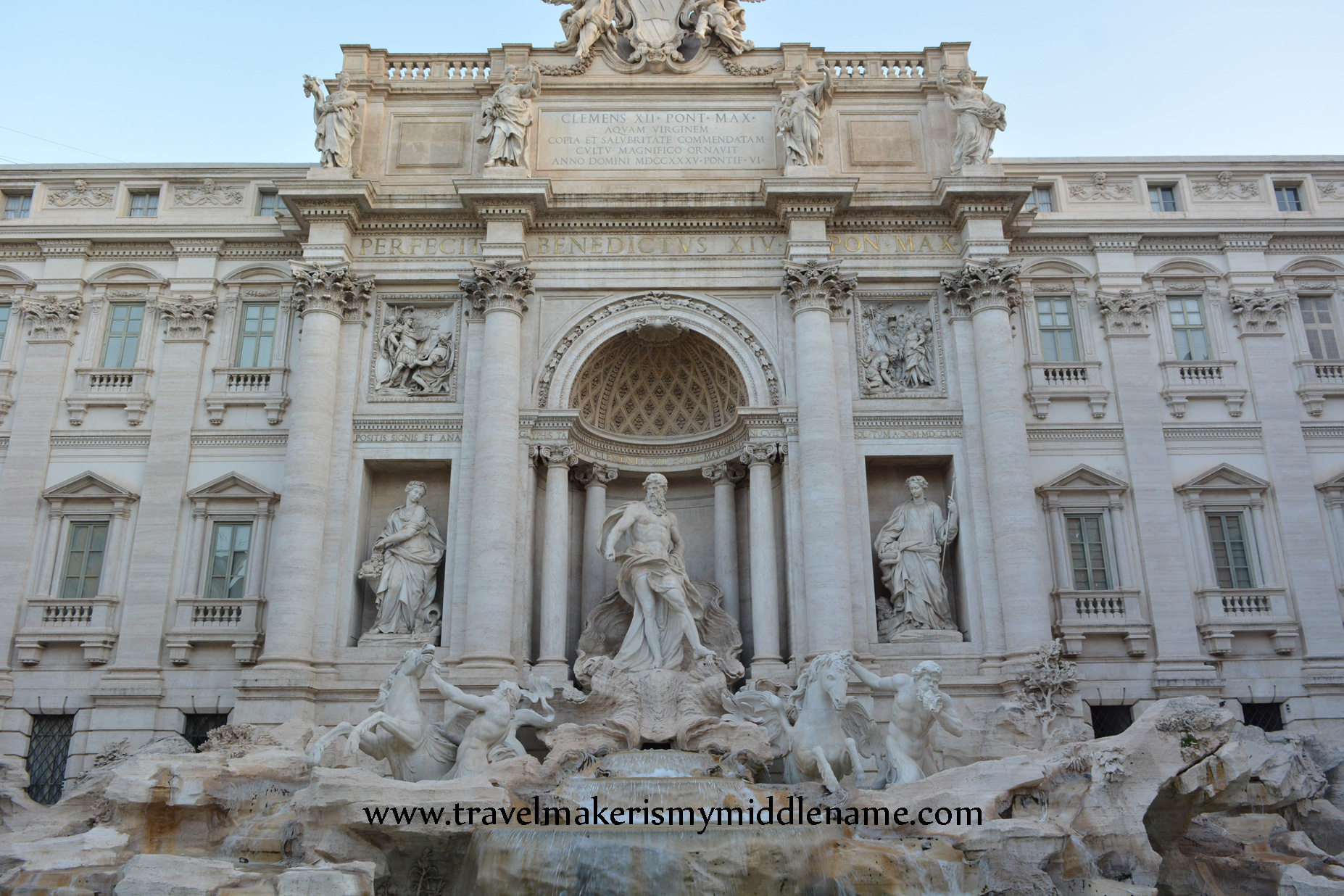 A front view of the Trevi Fountain during the day time with a blue cloudless sky in the top.