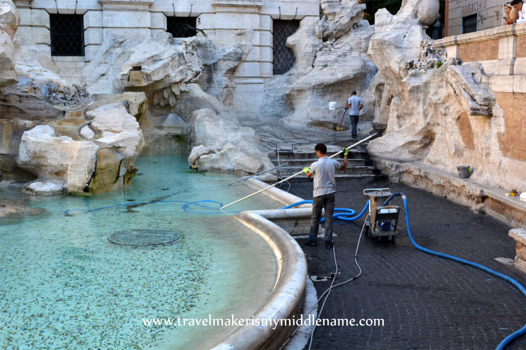 A man is fishing out coins in the Trevi Fountain basin and another man in the background sweeps the fountain.