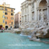 A side view of the Trevi Fountain showing the basin with blue water and water flowing into the basin from the sculptures during the day.