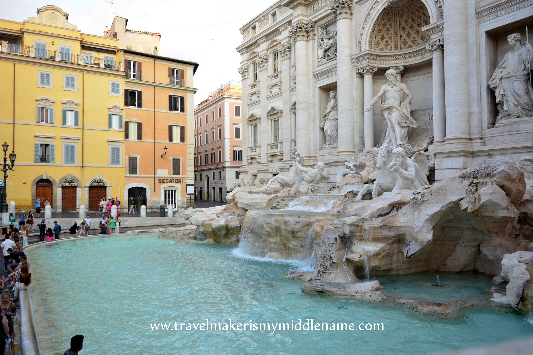 A side view of the Trevi Fountain showing the basin with blue water and water flowing into the basin from the sculptures during the day.