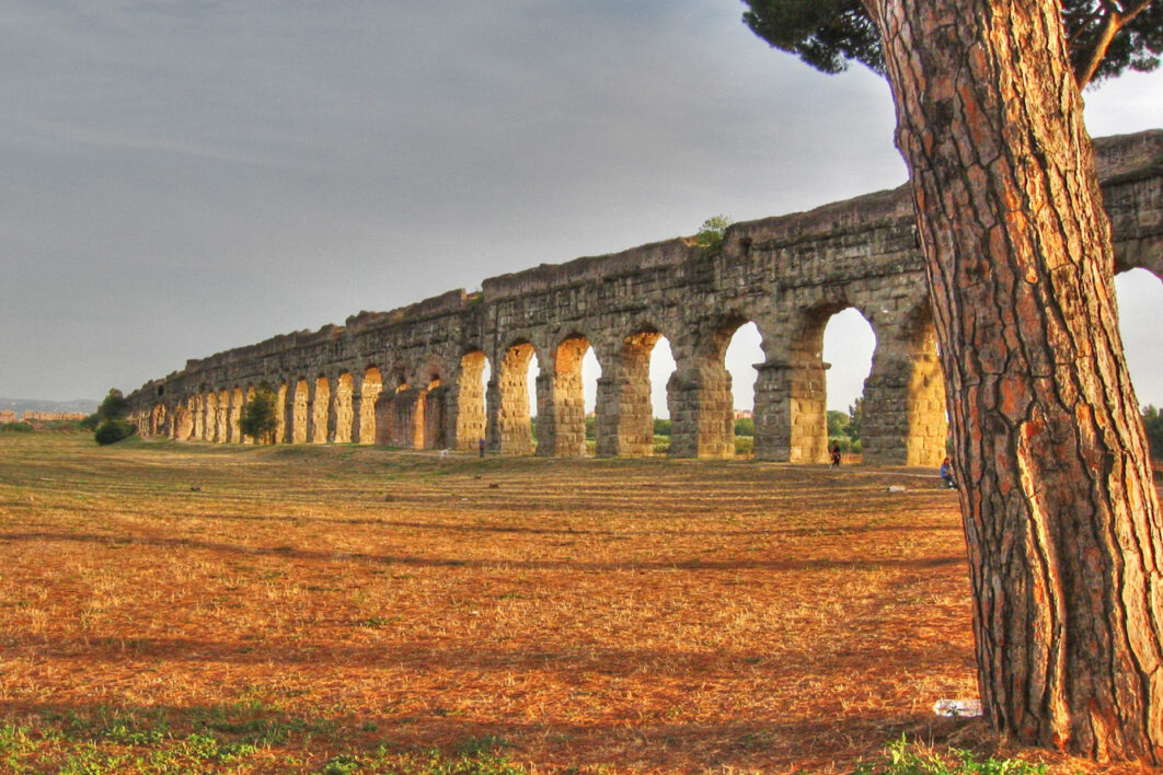 A Roman aqueduct stretches from the left to the right of the screen in a filed with a tree in the foreground on the right of frame.