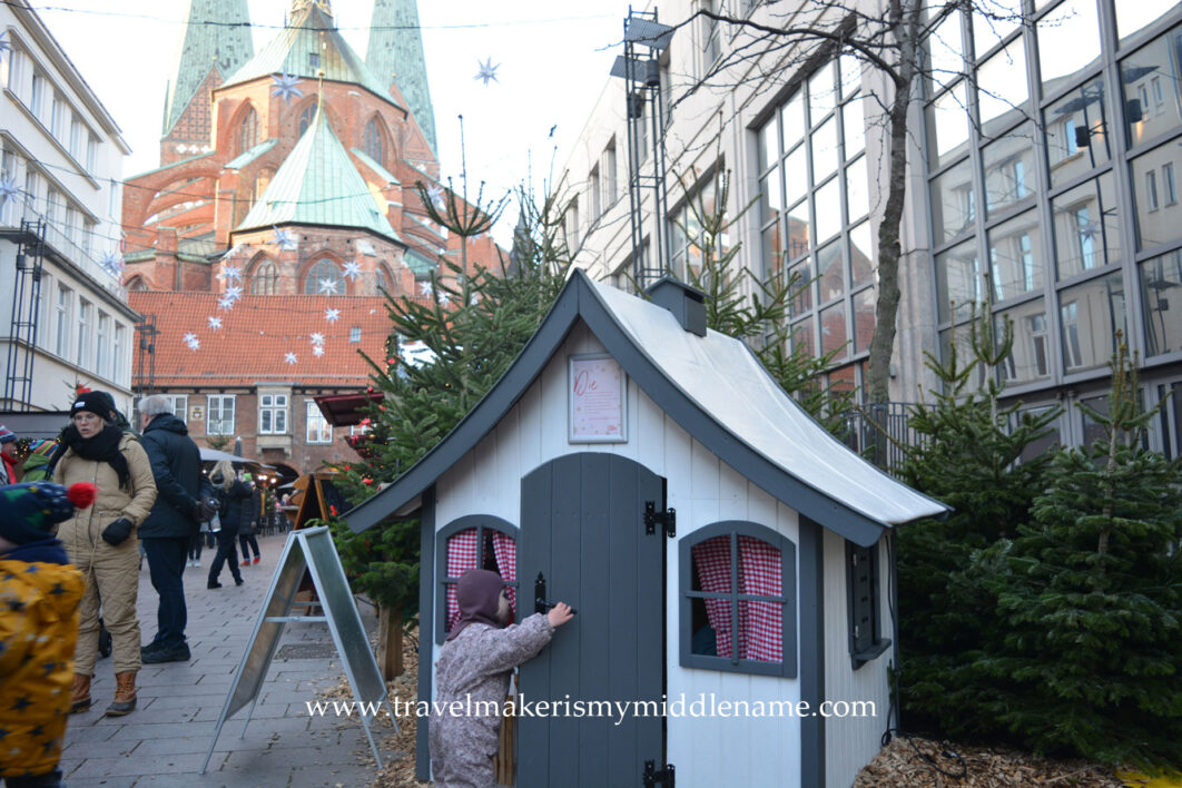 Something for everyone: Day time: A toddler at the Christmas market opens a door to a tiny house for Wichtel - a small gnome-like magical creature that appears around Christmas. In the background is the Marien Church of Lübeck with red brick and light green roof.