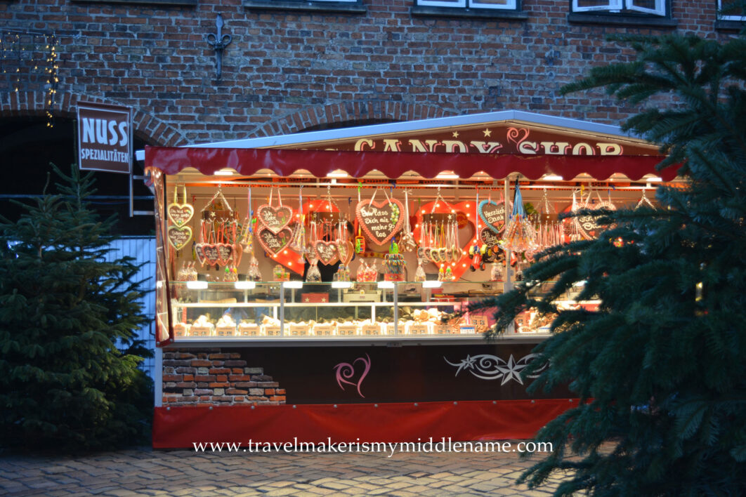 A large, brightly lit sweets and nuts stall at the Christmas market. On the right is a green Christmas tree, only half of it is in the frame. You can see the ground is covered in neatly paved cobblestones.