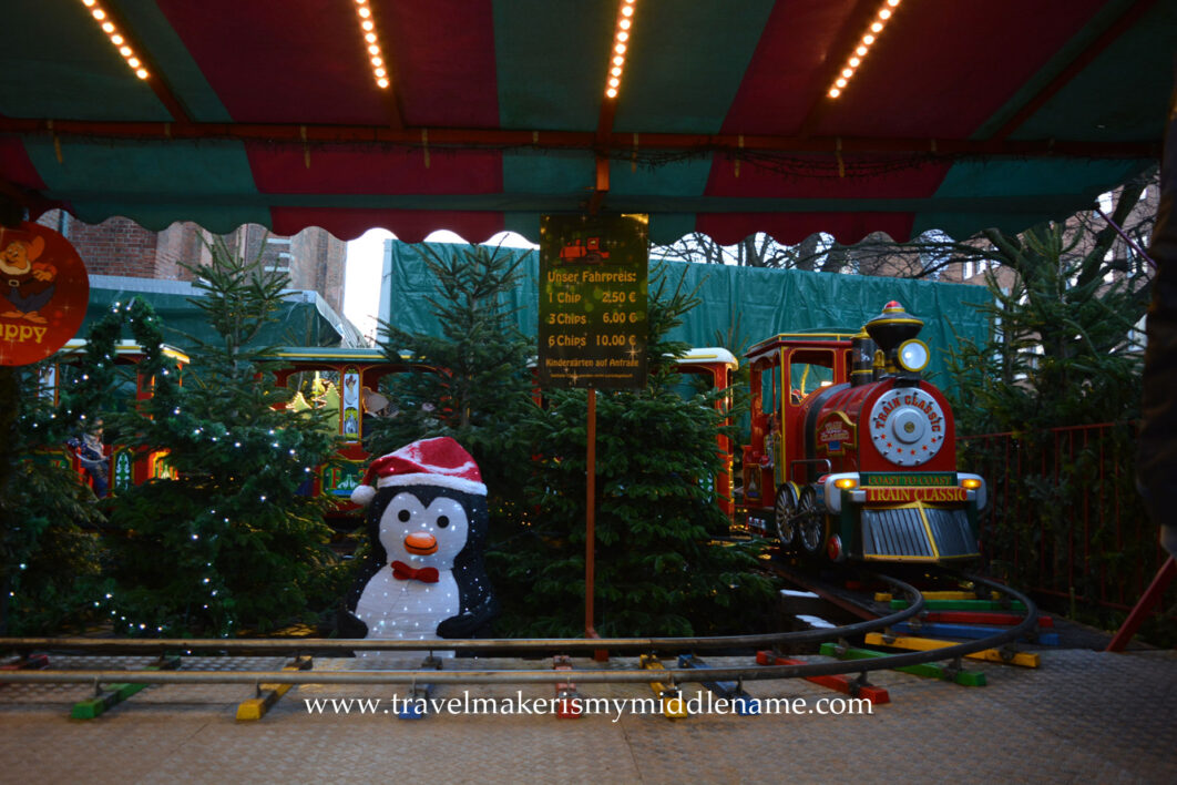Children (hidden behind the trees) ride on a colour train ride at the Christmas market in Lübeck. A penguin is in front.