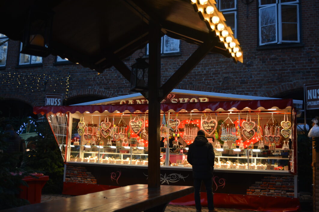 A man standing in front of a large, brightly lit sweets and nuts stall at the Christmas market pondering what to buy. In the foreground of the photo is a table with a roof where visitors can eat their food.