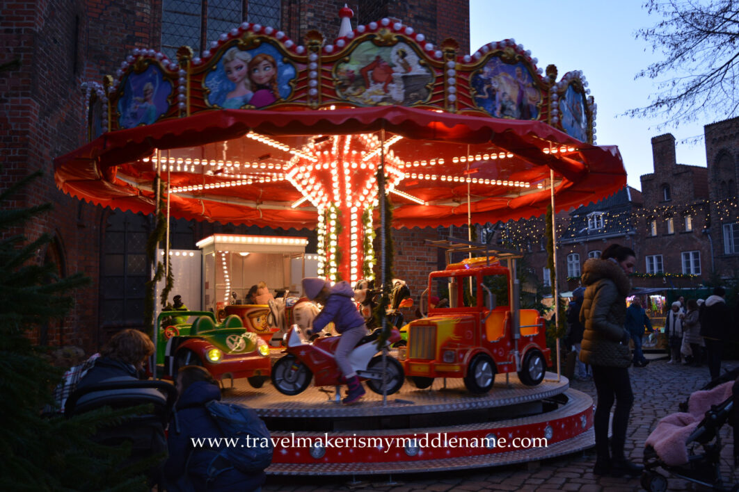 Something for everyone: A child riding on a motorcycle merry-go-round at the Christmas market. The merry-go-round has a red roof brightly lit with white light radiating from the center outwards.