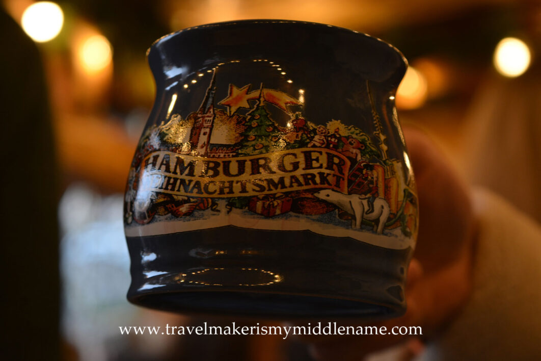 A ceramic mug with a painting of a Christmas market and the words "Hamburg Christmas market" on it in German