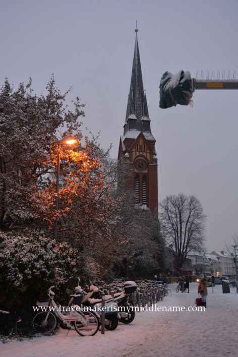 Street scene of Lübeck in winter in the evening when lights start to turn on.  A streetlight shines an reddish orange glow on some tree branches dusted in snow. On the ground is a row of rental bicycles covered in snow. In the background is the St. Lorenz Church.