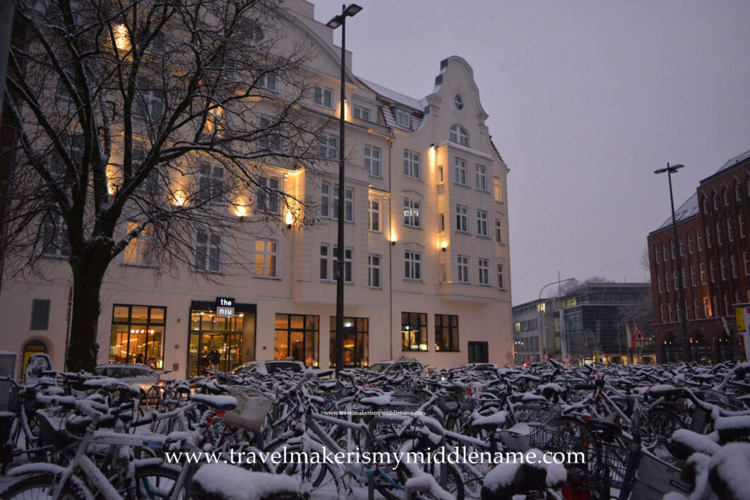 A building happily lit with welcoming yellow light in the background and rows upon rows of snow covered bicycles in the late afternoon in winter in Lübeck in mid December.