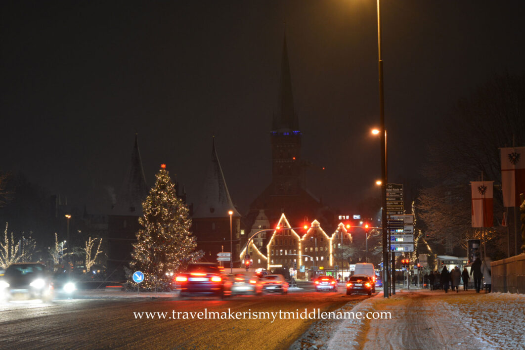 The streets of Lübeck with the Holsten Gate in the background to the left in the middle of December at night with snow covered footpaths on the right, and traffic on the road in the middle. A lit up Christmas tree is in the background to the left, and a building outline is lit up with lights.