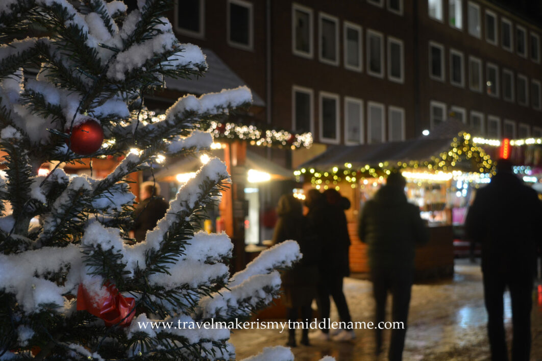 A small Christmas tree dusted with snow and red baubles on the left and people walking in the Christmas market in the background in Lübeck in the middle of December at night