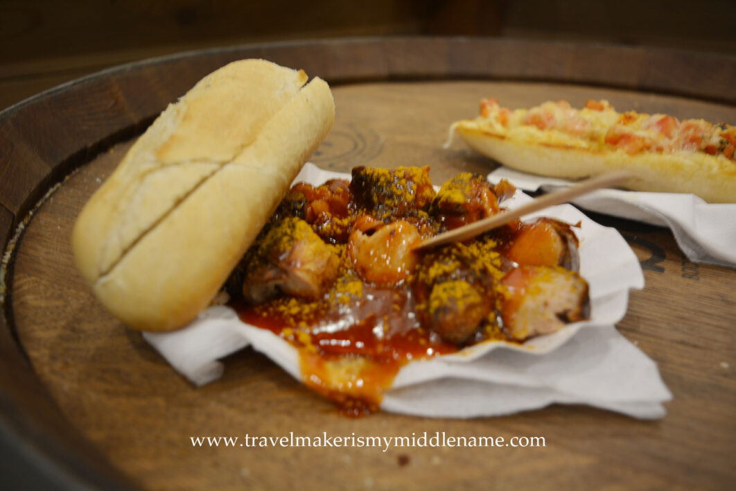 Chopped up pieces of sausages topped with sauce, served on a paper plate with a bread roll and toothpick.