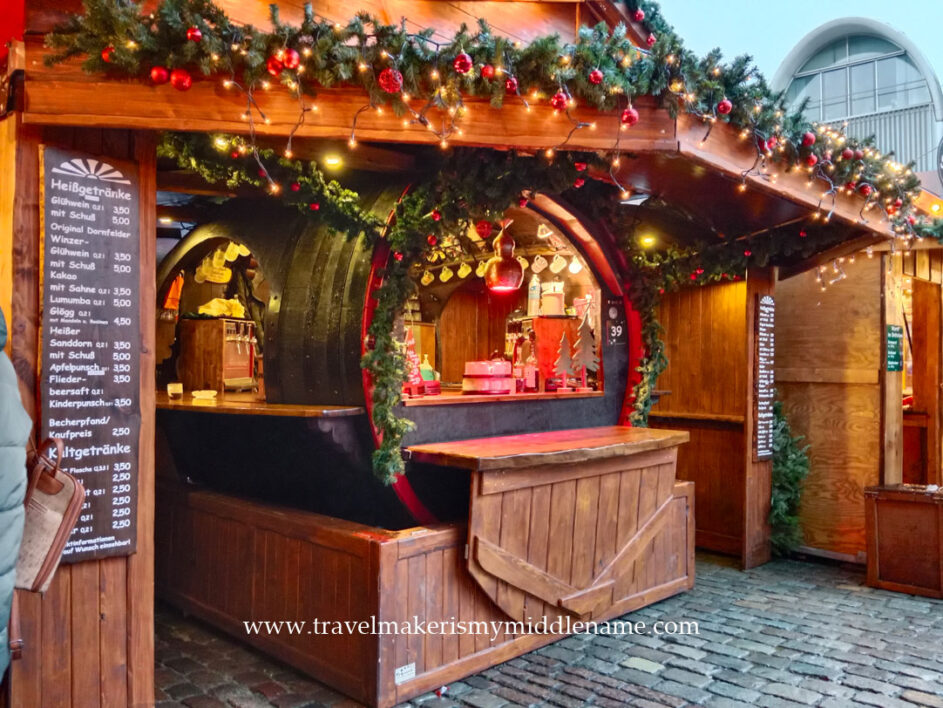A drinks stall in the shape of a large, black wine barrel on its side, inside a timbre hut. The roofline is decorated with tinsel, red baubles and lights