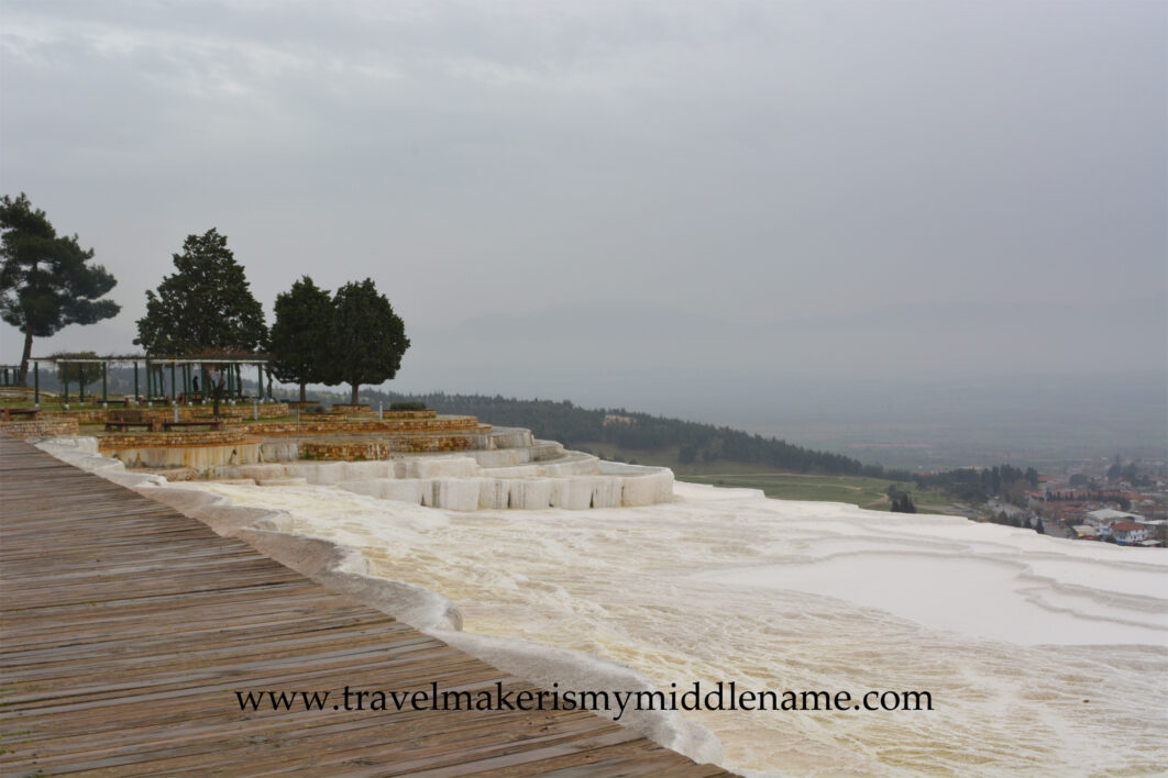 The white travertine terraces of Pamukkale in Turkiye, with a timber boardwalk around it on the left, trees in the distance, and a valley with buildings in the far distance.
