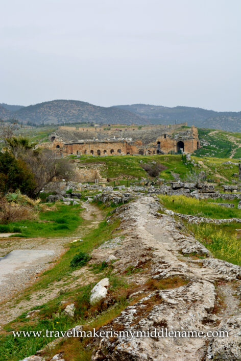 A cloudy day: A stone ridge among the hilly natural landscapes covered in green grass and dotted with small yellow flowers leads to the Hierapolis amphitheater in the distance in Pamukkale, Türkiye.