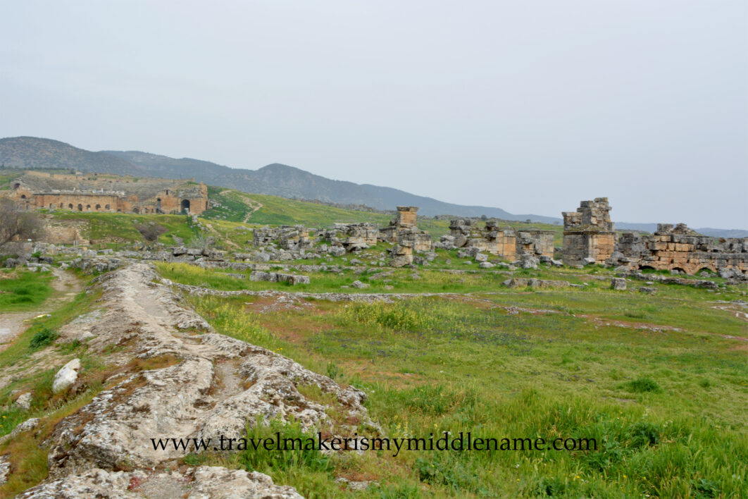 A cloudy day. A stone ridge among the hilly natural landscapes covered in green grass and dotted with small yellow flowers leads to the Hierapolis amphitheater in the distance to the left in Pamukkale in Turkiye. More stone ruins are on the right.