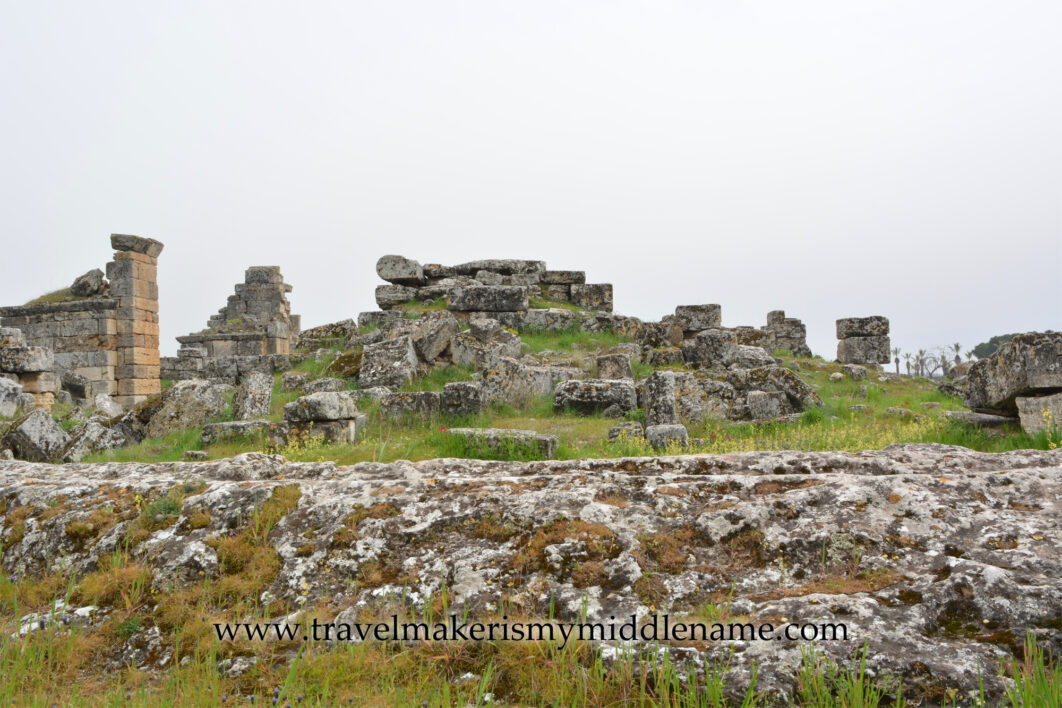 Some stone ruins in Hierapolis Pamukkale in Turkiye among the hilly natural landscapes covered in green grass in the background. Stones covered in brown moss in the foreground on a cloudy day.