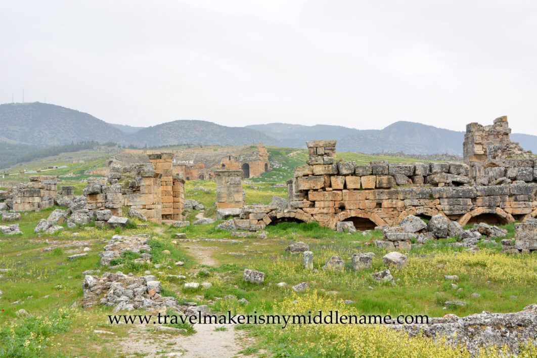 Some stone ruins in Hierapolis Pamukkale in Turkiye among the hilly natural landscapes covered in green grass and dotted with small yellow flowers on a cloudy day.