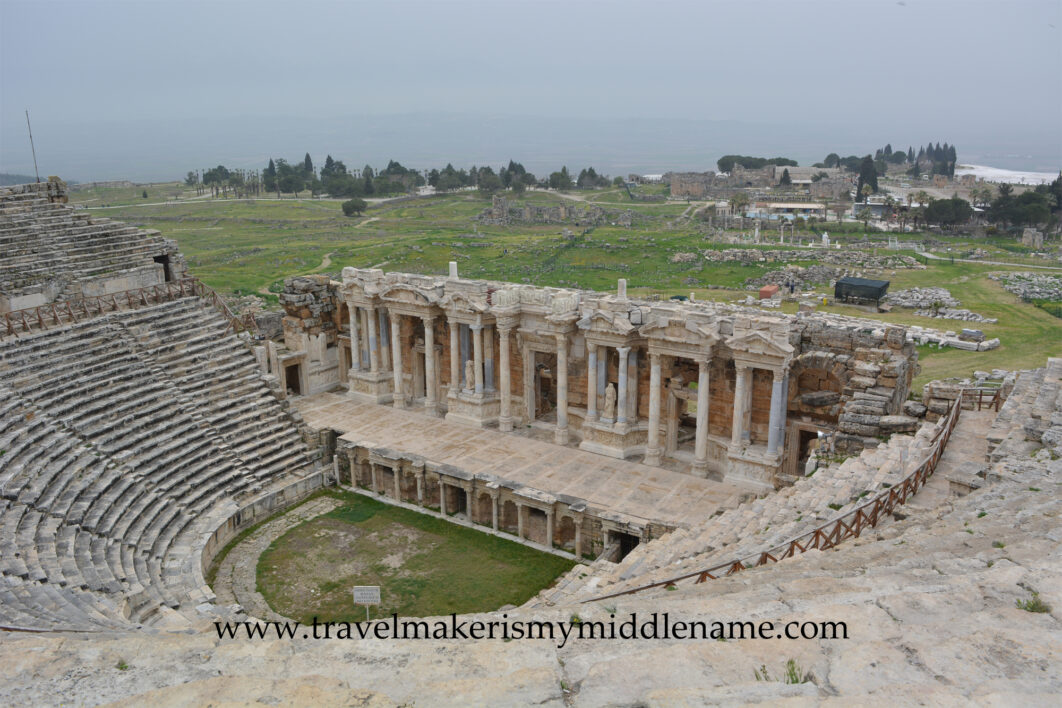 A view of the stone Hierapolis amphitheater on a cloudy day from the top to the bottom showing the stage and sculptures in the middle. The hilly natural landscapes covered in grass and trees in the distance in Pamukkale, Turkiye.