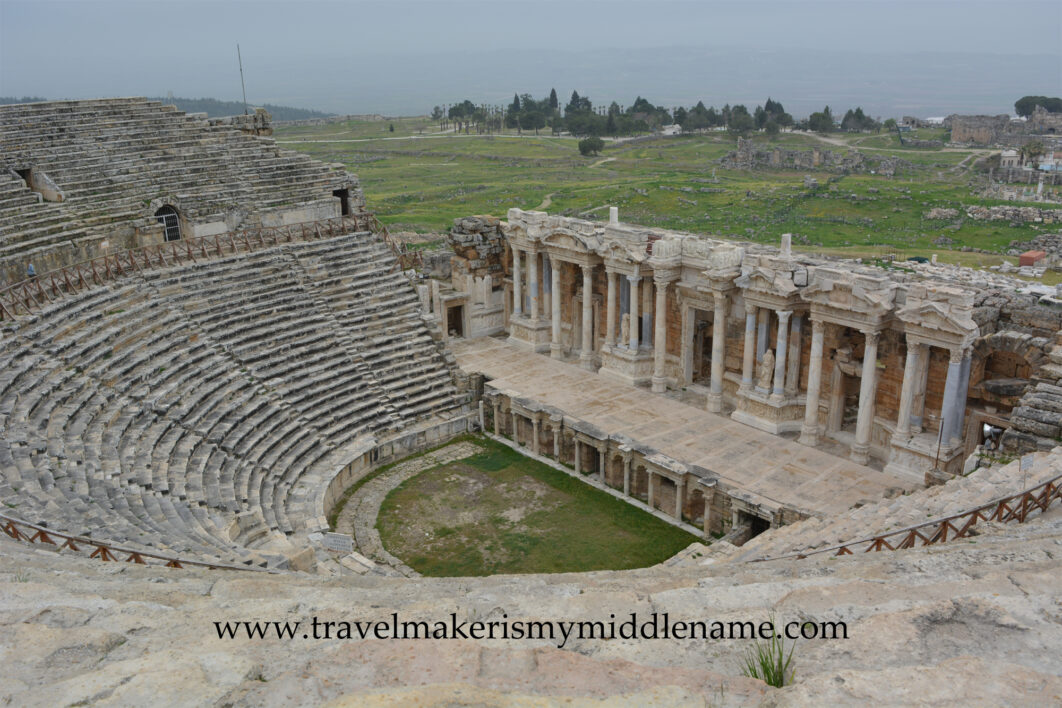 A view of the stone Hierapolis amphitheater on a cloudy day from the top to the bottom showing the spectator seats with the hilly natural landscapes covered in grass and trees in the distance in Pamukkale, Turkiye.