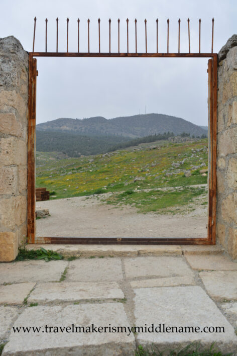 A cloudy day: A large rectangular doorway at the Hierapolis amphitheater at Pamukkale in Turkiye looking out to the hilly natural landscapes covered in green grass and dotted with small yellow flowers.