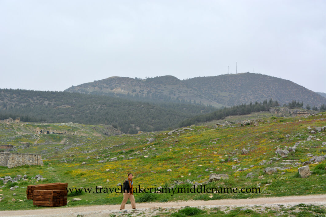 A cloudy day. A person walks up a dusty and rocky hill outside to the Hierapolis amphitheater in Hierapolis Pamukkale, Türkiye. In the background there is a natural landscapes covered in green grass and dotted with small yellow flowers.