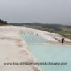 The white travertine terraces of Pamukkale on a cloudy day. Visitors walking in the light blue coloured pool and the white travertine formations around it. The city can be seen in the distance.