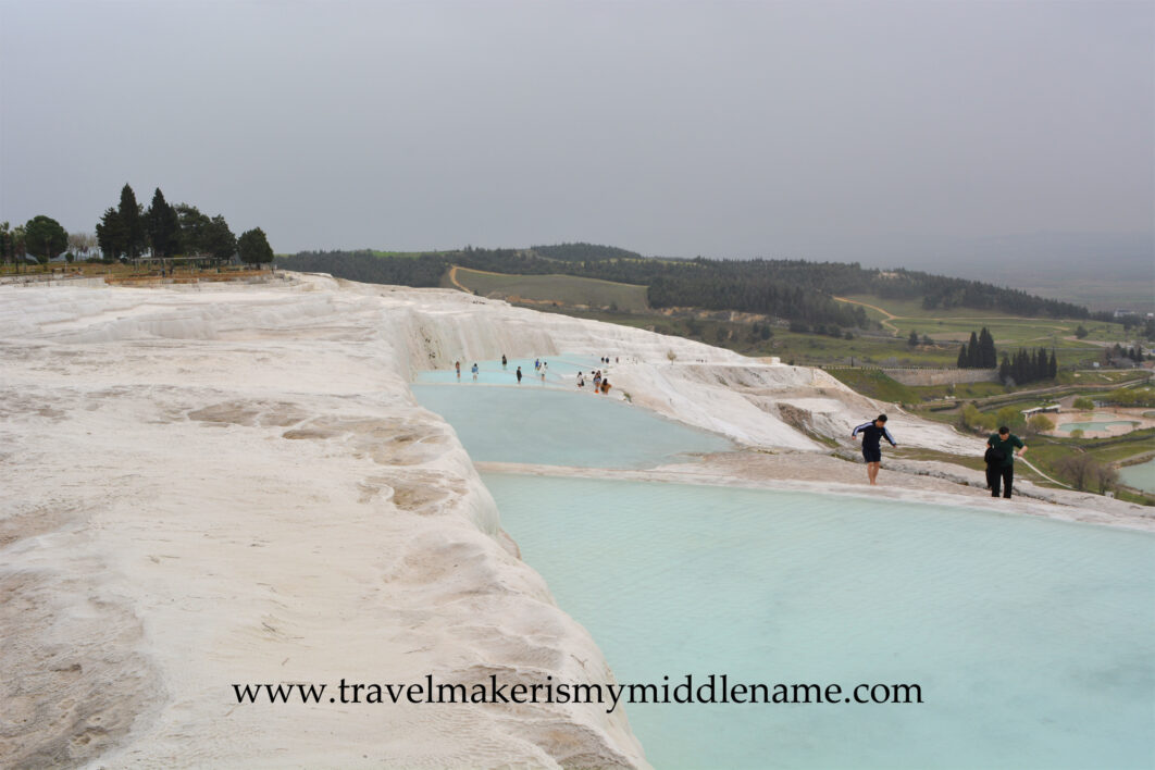 The white travertine terraces of Pamukkale on a cloudy day. Visitors walking in the light blue coloured pool and the white travertine formations around it. The city can be seen in the distance.