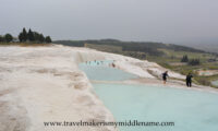 The white travertine terraces of Pamukkale on a cloudy day. Visitors walking in the light blue coloured pool and the white travertine formations around it. The city can be seen in the distance.