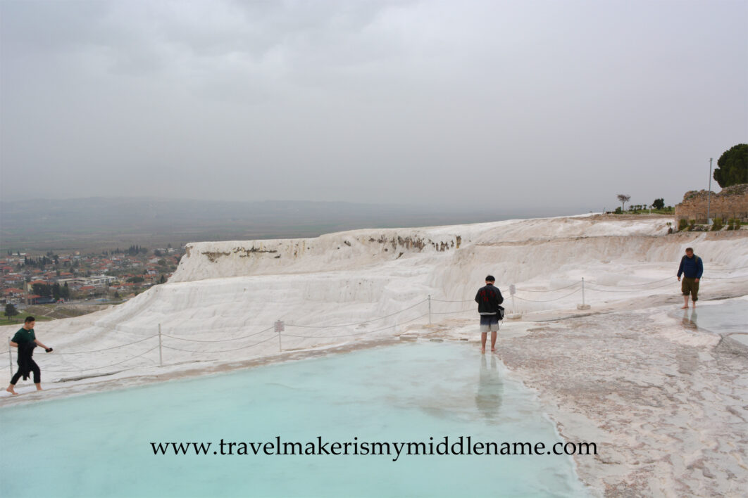 The white travertine terraces of Pamukkale on a cloudy day. Some people walk along the railing of the light blue coloured pool where it meets the white travertine formations around it. The city can be seen in the distance.