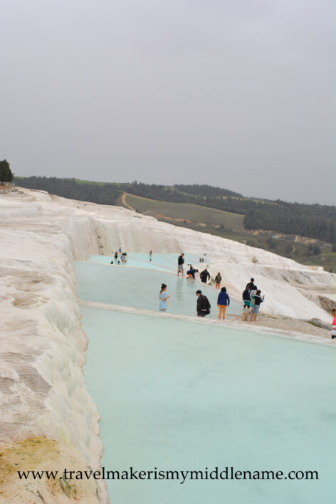 The white travertine terraces of Pamukkale on a cloudy day. A portrait oriented photo showing visitors walking in the light blue coloured pool and the white travertine formations around it. The city can be seen in the distance.