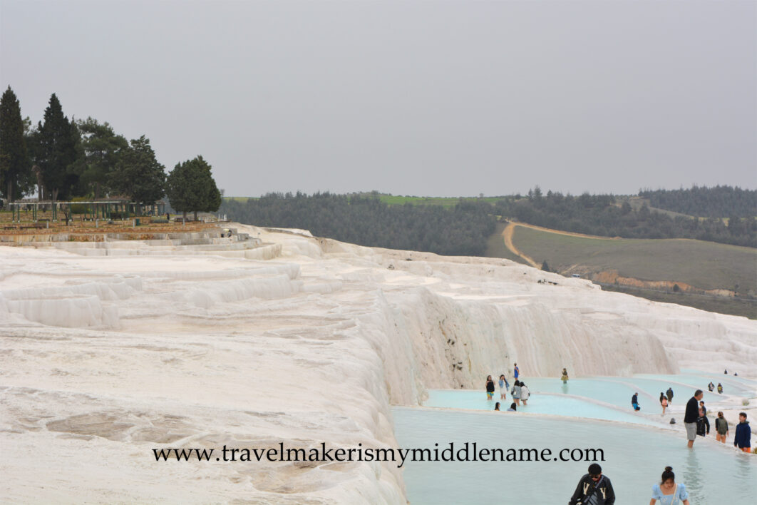 The white travertine terraces of Pamukkale on a cloudy day. Visitors walk in the pool on the right side of the frame. The city can be seen in the distance.