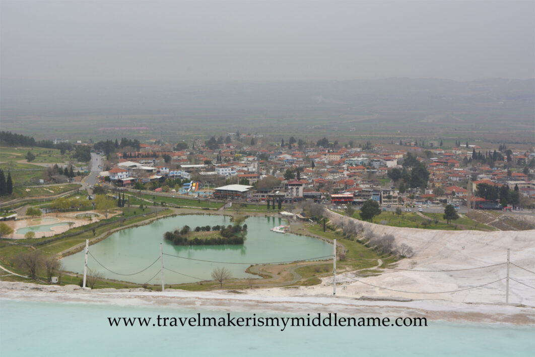 The white travertine terraces of Pamukkale surrounding a pool in a lower region and the city below in the distance on a cloudy day.