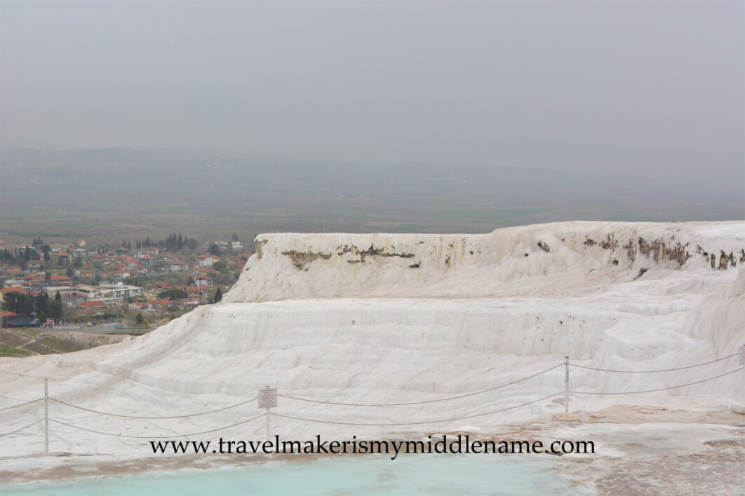 The white travertine terraces of Pamukkale, Türkiye, showing the hardened calcium deposits dripping downwards, with the city in the valley below in the distance on a cloudy day.
