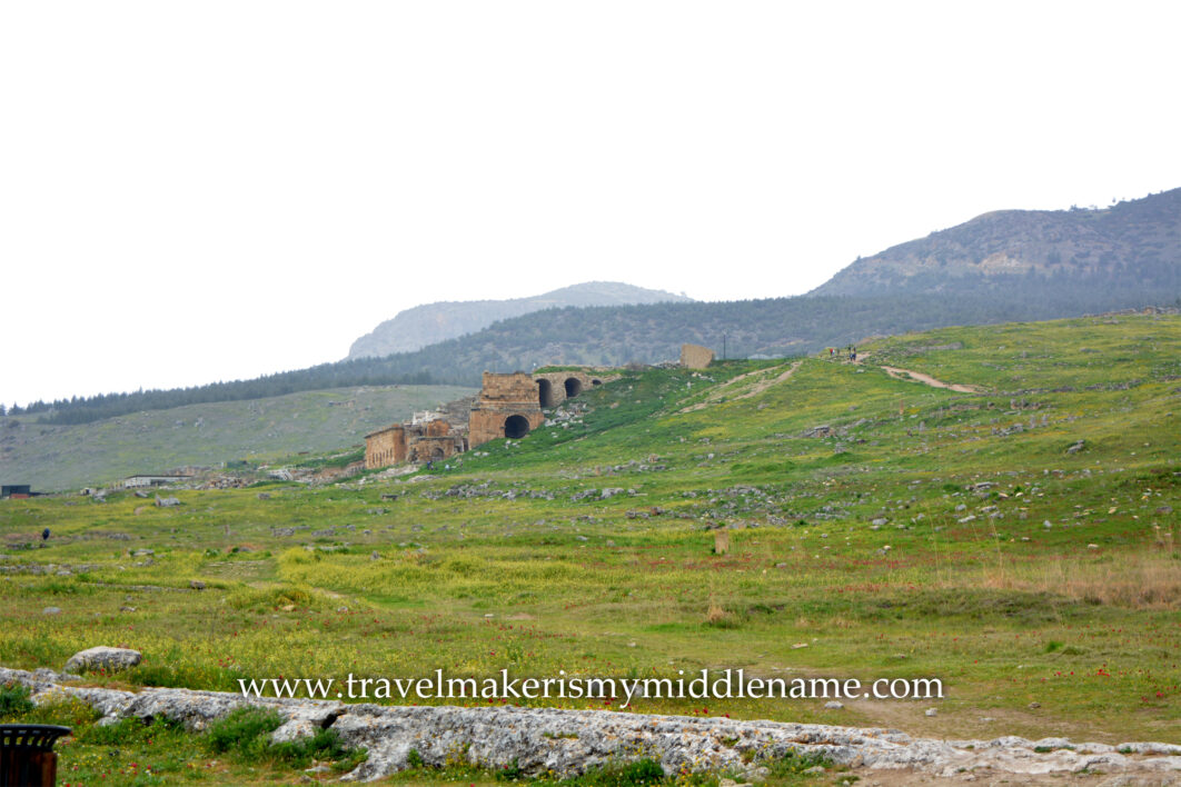 A cloudy day. Ruins of a stone bridge-like structure with arches underneath in the background among the hilly natural landscapes covered in green grass in Pamukkale in Turkiye.