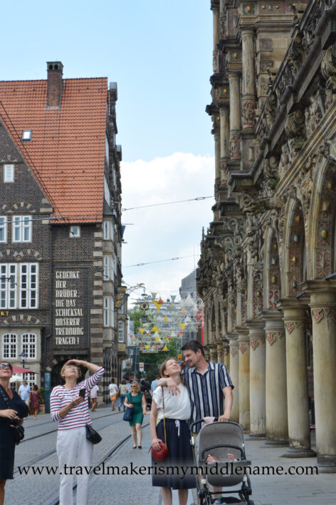 A portrait oriented photo showing people walking in the street outside the Bremen marketplace, showing half of a brick building on the left and the stone columns of the Bremen Rathaus on the right. A woman on the left hand side looks upwards with her left hand on her head. A couple in the middle walks together. The man is pushing a stroller. His right arm is around the woman, who he is looking at. The woman is in a white shirt and navy skirt, and has a small red bag.