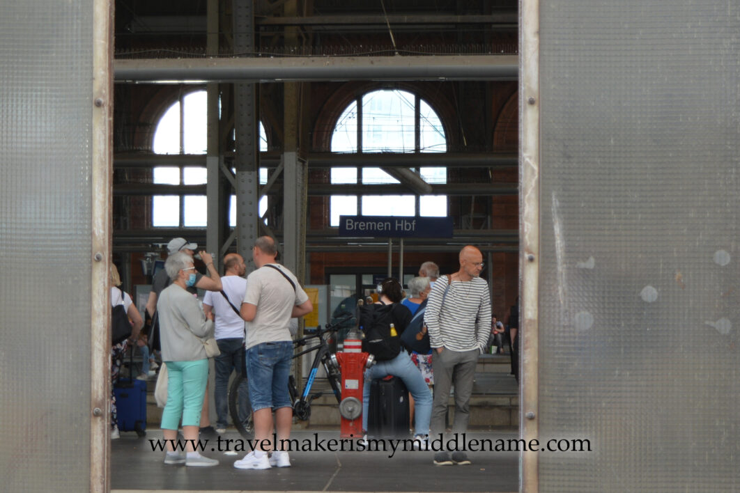 People on the train station at Bremen central train station as seen through a wide gap in a metal screen. 