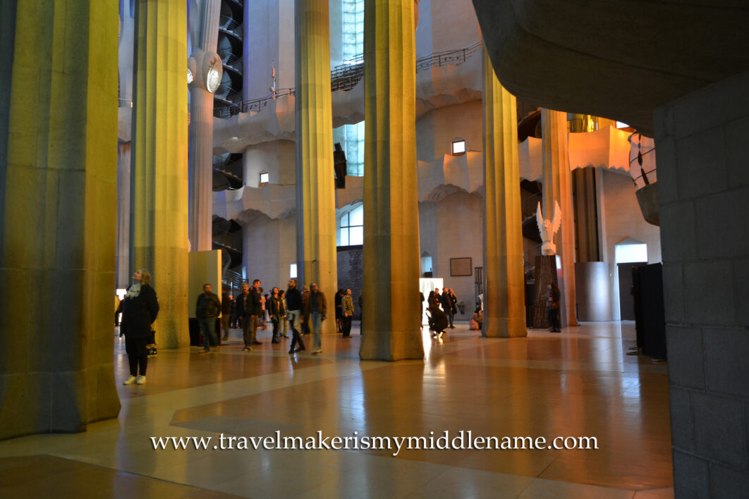 A wide shot of people walking through the many columns inside La Sagrada Familia church in Barcelona, Spain at ground level inside. Orange afternoon light reflects off the columns. The high celing is out of frame. 