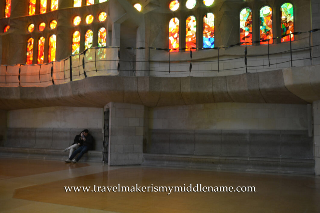 Afternoon sunlight shines through the red, yellow, and orange vertical stained glass windows inside La Sagrada Familia church in Barcelona, Spain. Two people sit on some seats beneath the balcony that are under the stained glass windows.
