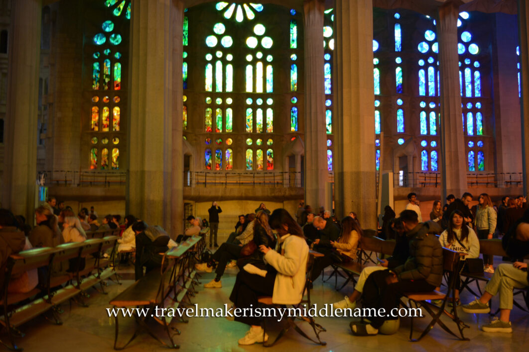 Side view of visitors facing the left of the frame bathed in afternoon orange sun, resting in the spacious pews on the ground floor of La Sagrada Familia in Barcelona, Spain. The background is a wall with colourful stained glass windows and two stone columns in the centre.