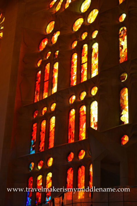 Afternoon sunlight shines through the red, yellow, and orange vertical stained glass windows at ground level inside La Sagrada Familia church in Barcelona, Spain.
