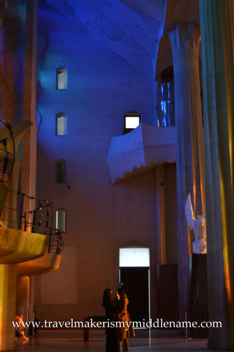 Two girls taking photos of the view on the ground floor inside La Sagrada Familia in Barcelona, Spain on a sunny afternoon. The cavernous ceiling, bathed in blue light, towers over them. The interior walls and balcony are bathed in orange light. 