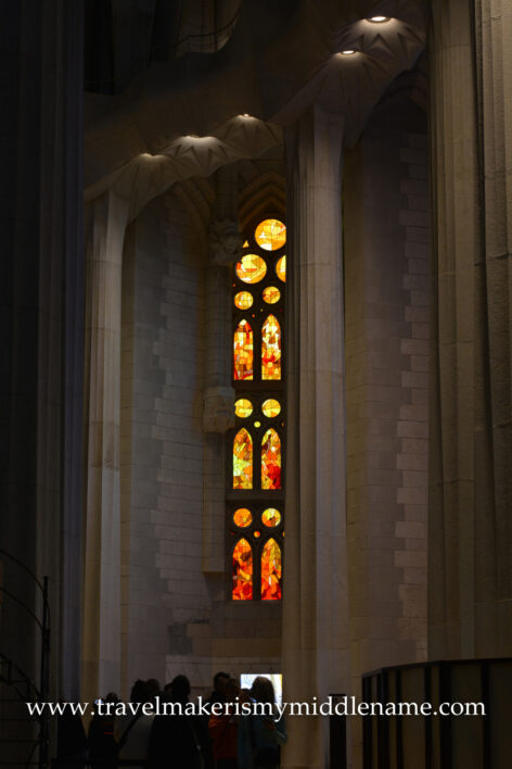 A vertical sliver of stained glass window behind some columns and stone walls in La Sagrada Familia in Barcelona, Spain. 
