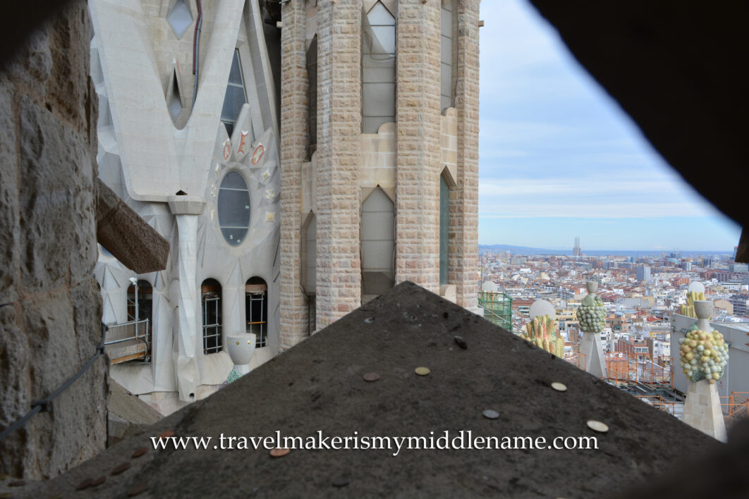 A triangular stone structure pointed outwards towards a view of the side of a tower La Sagrada Famiila church in Barcelona, Spain, at the tower height. The triangular stone structure guides rain away from the window below. People throw coins on to the ledge for fun. Yellow and green fruit sculptures can be seen in the bottom right corner.