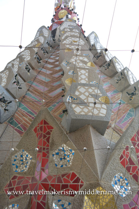 A view of the colourful red, pink, and yellow tile mosaic facade and the peak of the tower in La Sagrada Familia church in Barcelona, Spain, as seen from the Passion tower bridge between the tower at around 90m.
